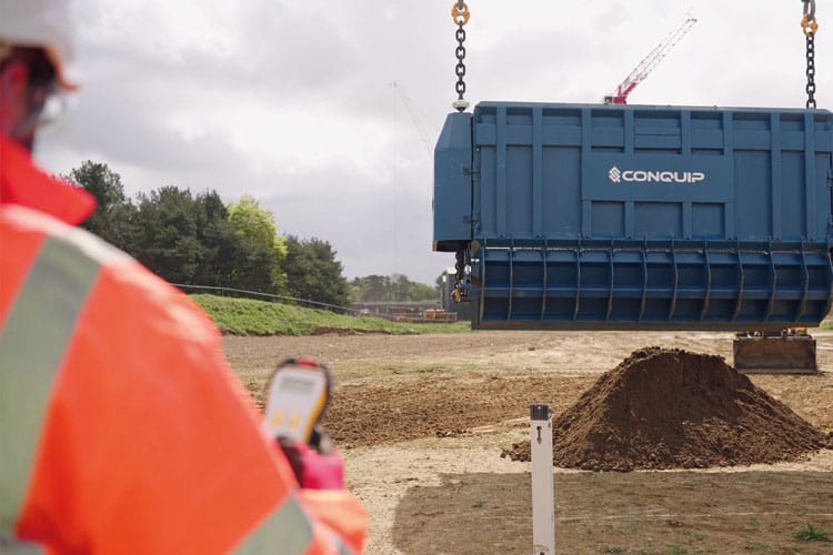 A worker in an orange safety jacket operates a remote control, guiding a large blue Conquip Smart-Release BulkX being lifted by a crane. The background features a grassy landscape and construction equipment under a cloudy sky.